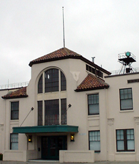 The facade of Hangar 5 is cream-colored. The entrance is at the base of a short tower, which has an arched window, tile roofing, and a flag pole. There is a blue beacon to the right of the tower.