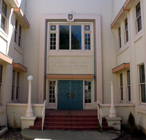 The old airport hotel is a two-story pastel pink building with teal and light red trim. Stairs lead to the entrance. There are decorative windows beside and above the entrance, and ordinary windows on the side walls that enclose the entrance courtyard. Lampposts flank the entrance. The right lamppost is missing its glass globe.