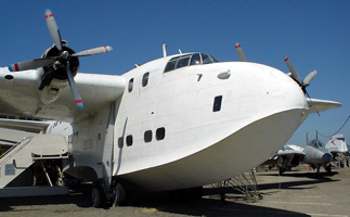 The Solent flying boat is white. One propeller is visible on the left side of the picture; two are visible on the right. The wings meet the plane at the top of fuselage, not far behind the windshield, which has a bubble shape. There are small, squarish windows at various levels along the fuselage.